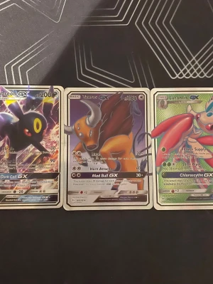 Pokémon GX-kort Umbreon, Tauros, Lurantis - Tre Pokémon GX-samlarkort: Umbreon GX i svart med lila och gula detaljer, Tauros GX i brunt och orange samt Lurantis GX i grönt och rött. Alla kort har holografisk yta och klassisk rektangulär form. Perfekt för Pokémon-fans och samlare.