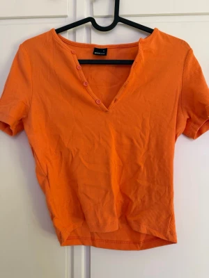 Orange ribbad topp från Gina Tricot - Kortärmad orange topp från Gina Tricot i ribbad bomull med v-ringning och dekorativa knappar framtill. Modellen är croppad och har en enkel, stilren look som passar perfekt till jeans eller kjol.