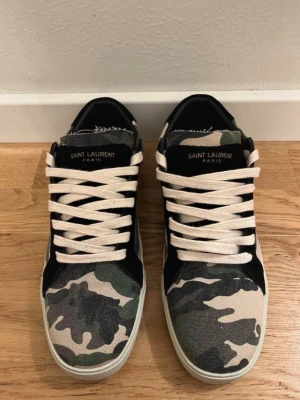 Saint Laurent sneakers med camomönster - Säljer ett par Saint Laurent sneakers med grymt camomönster i grönt, svart och beige. Skorna har vita snören, svart detalj vid snörningen och tydlig Saint Laurent-logga på sidan och plösen. Ovandelen är i canvas och sulan är vit och platt.