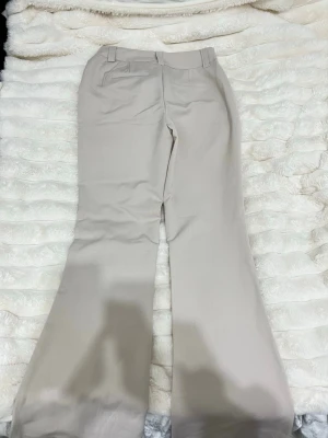 Beiga bootcut kostymbyxor - Snygga beiga kostymbyxor med bootcut passform och hög midja. Byxorna har klassiska fickor bak och framtill samt bälteshällor i midjan. Perfekta för en clean och stilren look. Materialet känns som en blandning av bomull och polyester.