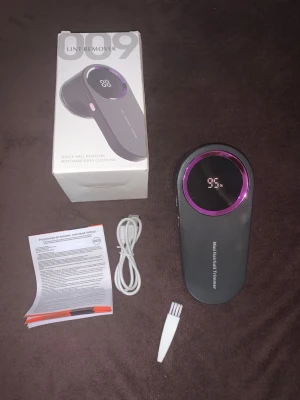 Elektrisk ludd- och nopprengörare - Elektrisk ludd- och nopprengörare i svart plast med lila detaljer och digital display. Levereras med USB-laddkabel, rengöringsborste och manual. Perfekt för att ta bort noppor från kläder och textilier.