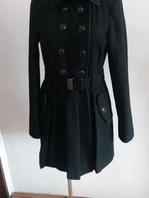 Svart trenchcoat med bälte bh - Stilren svart trenchcoat med dubbelknäppning och bälte i midjan. Klassisk krage, långa ärmar och fickor med knappdetaljer. Perfekt för dig som vill ha en tidlös och snygg jacka till höst och vinter.