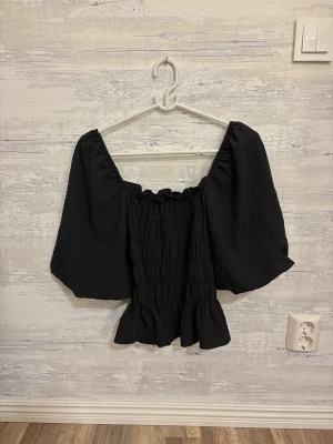 Svart topp med puffärm - Trendig svart topp med breda puffärmar och smockad kropp. Toppen har en volangkant nedtill och är tillverkad i ett lätt, strukturerat tyg som ger en snygg siluett. Perfekt för dig som vill ha en statement topp till din outfit.