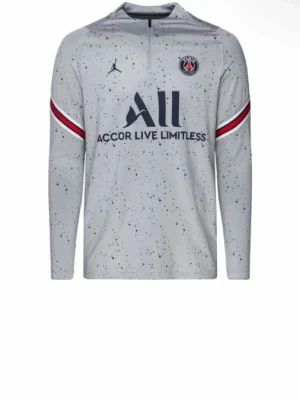 Grå PSG träningströja Jordan - Snygg grå långärmad träningströja från PSG och Jordan med blåa prickar, halv dragkedja och klubbmärke på bröstet. Röd och blå rand på ärmarna och stor logga framtill. Perfekt för fotbollsträning eller chill.