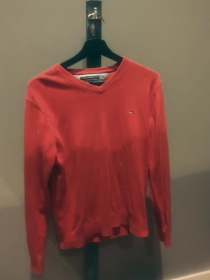 Röd V-ringad tröja Tommy Hilfiger - Röd stickad tröja från Tommy Hilfiger med V-ringning och liten logga broderad på bröstet. Tröjan har långa ärmar och ribbade muddar vid ärmslut och nederkant. Perfekt för dig som gillar klassisk och stilren design.
