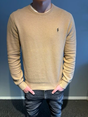 Ralph Lauren tröja  - Snygg beige stickad tröja från Polo Ralph Lauren med rund halsringning och diskret broderad logga på bröstet. Tröjan har en klassisk passform. Tillverkad i mjukt material som känns skönt mot huden. Storlek M. Modellen är 184cm.