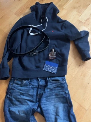 Mörkblå hoodie från Polo Ralph Lauren - Mörkblå hoodie från Polo Ralph Lauren med klassisk huva och vita dragsnören. Liten röd logga broderad på bröstet och stor känguruficka framtill. Tillverkad i mjuk bomullsblandning, perfekt för en avslappnad och stilren look.