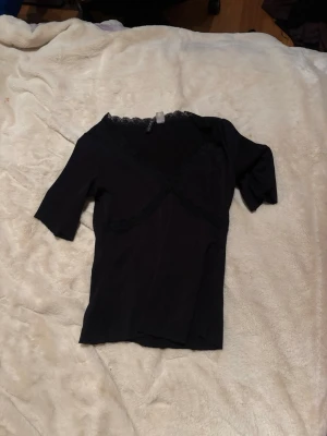 Svart topp med spetsdetalj - Snygg svart topp med v-ringning och spetsdetaljer längs halsringningen. Modellen har korta ärmar och en figurnära passform som framhäver siluetten. Perfekt att styla med jeans eller kjol för en trendig look.