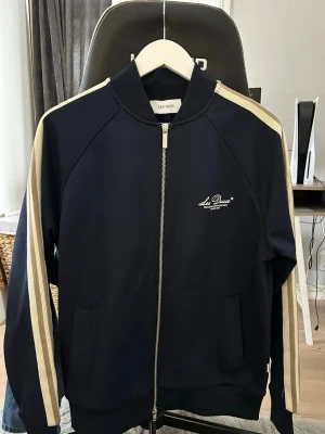 Mörkblå ziptröja Les Deux - Snygg mörkblå ziptröja från Les Deux med vita ränder längs ärmarna och logga på bröstet. Tröjan har hög krage, dragkedja framtill och två fickor. Perfekt för en sportig och avslappnad stil. Ordinarie pris 1500