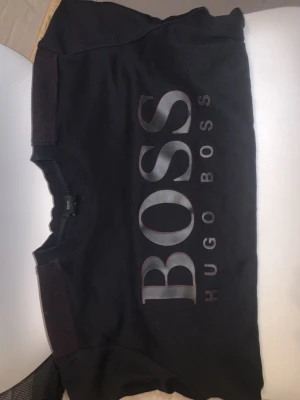 Svart sweatshirt från Hugo Boss - Svart sweatshirt med stor BOSS-logga och texten HUGO BOSS på bröstet. Tröjan har rund halsringning och ribbade muddar vid ärmslut och nederkant. Perfekt för en stilren och avslappnad look.