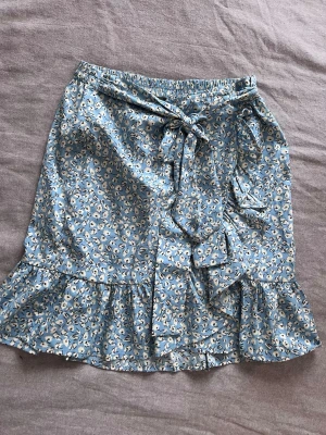 Blommig blå omlottkjol med volang - Söt kort omlottkjol i ljusblått med små vita blommor och volangkant. Kjolen har knyt i midjan och elastisk resår för skön passform. Perfekt för vår och sommar när du vill ha en lätt och luftig look. 