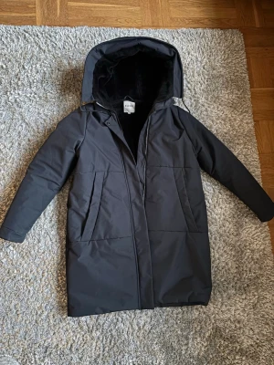 Jacka ELVINE storlek L - Mörkblå vinterjacka från ELVINE. Storlek L. Använd något år men är i gott skick. Lite slitake runt ärmarna (se bild). Rak modell och fuskpälsfodrad luva. Jackan är fodrad med Thermore som har samma egenskaper som dun. Den är vattentät och klarar vind. Medellång, varmfodrade fickor, dragkedja och dubbel knappslå med magnetknappar. Tror att modellen heter Tiril. Normal i storleken.