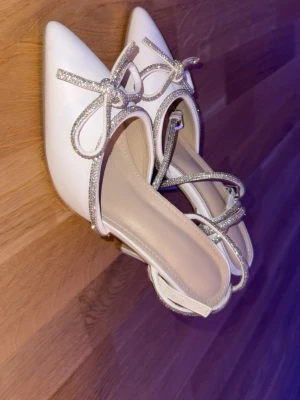 Vita pumps med glitterdetaljer och rosett - Snygga vita pumps med spetsig tå, smala remmar med glittriga detaljer och en söt rosett framtill. Skorna har hög klack och öppen häl, perfekta för att lyfta din outfit. Materialet ser ut att vara syntet med glitterinslag.