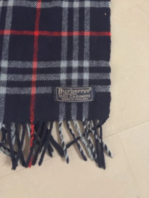 Rutig halsduk i kashmir från Burberrys - Mörkblå halsduk i 100% kashmir från Burberrys med klassiskt rutmönster i grått, vitt och rött. Halsduken har fransar i ändarna och en mjuk, lyxig känsla. Perfekt accessoar för att lyfta din outfit med en tidlös brittisk stil.