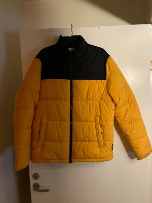 Gul och svart pufferjacka - Snygg pufferjacka i gul och svart med hög krage och quiltad design. Jackan har dragkedja framtill och två sidofickor. Perfekt för kalla dagar när du vill sticka ut lite extra.