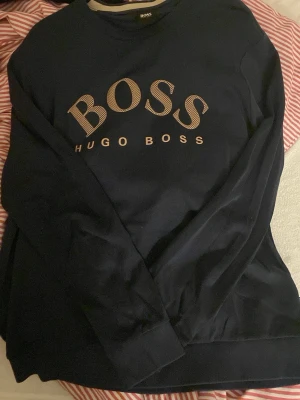 Mörkblå sweatshirt från Hugo Boss - Snygg mörkblå sweatshirt från Hugo Boss med stort BOSS-tryck i beige på bröstet. Tröjan har rund halsringning och långa ärmar. Storlek XL men sitter som L