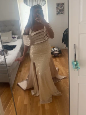 Champagnefärgad festklänning  - Elegant långklänning i champagnefärg med glittrig framsida, offshoulder-detalj och breda axelband. Klänningen har en hög slits framtill och ett långt släp baktill. Perfekt för bal eller festliga tillfällen. Materialet är glittrig polyester och satinliknande tyg. Ny pris 6000