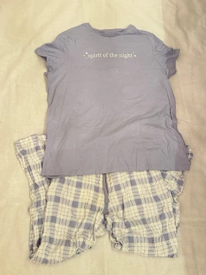 Lila pyjamas med text och rutiga byxor set  - Mysig pyjamas med lila t-shirt där det står 'spirit of the night' och matchande byxor i lila och vitt rutmönster. T-shirten har rund hals och korta ärmar, shortsen har avslappnad passform. Perfekt för chillkvällar hemma. I storlek L.