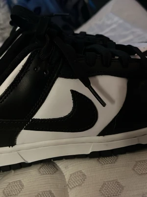 Nike Dunk Low svartvita sneakers - Säljer ett par klassiska Nike Dunk Low sneakers i svart och vitt. Skorna har en låg silhuett, snörning och den ikoniska Swoosh-loggan på sidan. Tillverkade i skinn med perforerad tå för extra ventilation. Perfekta för dig som gillar stilrena och tidlösa sneakers. Andvända 2 gånger 