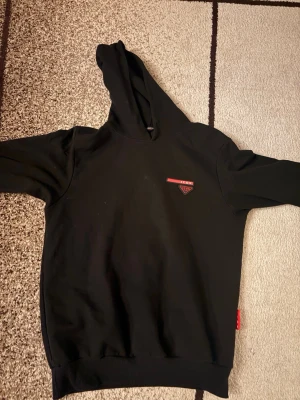 Svart hoodie från ICON med röd logga - Snygg svart hoodie från ICON med luva.  På högra bröst sidan  sitter en röd och svart logga med texten ICON. Hoodien är jätte skön och värmer upp dig jättesnabbt perfekt för vinter den är helt ny  men blivit liten för mig.       Min kamera tog  inte så bra bilder men verkligheten så är det helt ny 