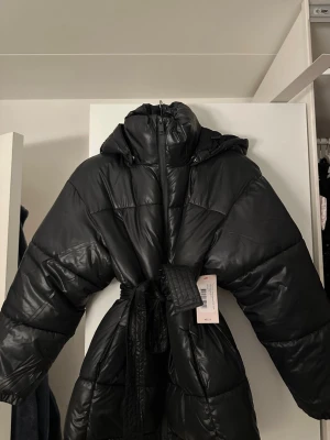 Svart pufferjacka med bälte och huva - Snygg svart pufferjacka med huva och bälte i midjan för en cool och oversized look. Jackan har quiltad design och dragkedja framtill. Perfekt för kalla dagar när du vill vara både varm och trendig. Den är till slutet av magen/början av låren. Helt ny med prislapp ! 🥰🩷