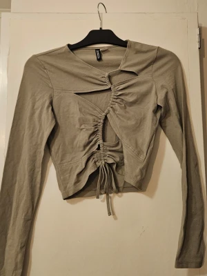 Grågrö  långärmad topp med snörning - Trendig grågrön långärmad topp från H&M med unik snörningsdetalj framtill och asymmetrisk design. Toppen är croppad och har coola cut-outs som ger en edgy look. Perfekt för dig som vill sticka ut med din stil.