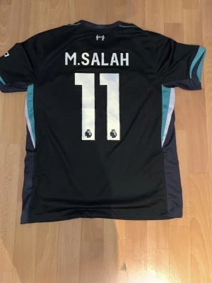 Liveepool Salah tröja 24/25 - Snygg svart Liverpool FC fotbollströja från Nike med M. Salah och nummer 11 på ryggen. Tröjan har vita och turkosa detaljer, klubbmärke och sponsorlogga framtill. Perfekt för fotbollsfans. Storlek M och väldigt nytt skick.