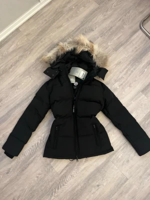 Chelsea parka Canada goose - Snygg svart dunjacka från Canada Goose med figurnära passform, huva med fuskpäls och dragkedja framtill. Jackan har två framfickor med dragkedja och klassisk logga på ärmen. Perfekt för kalla vinterdagar.