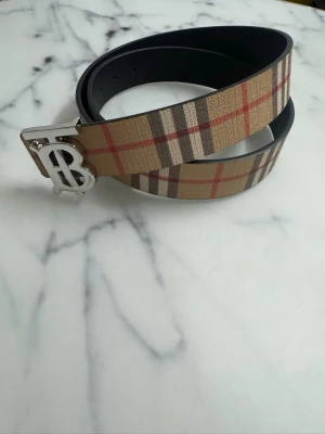 Burberry bälte  - Snyggt bälte från Burberry med det ikoniska beige, röda och svarta rutmönstret. Bältet har ett stort silverfärgat spänne med Burberrys TB-logga. Tillverkat i läder med canvasdetaljer. Perfekt accessoar för att lyfta din outfit.