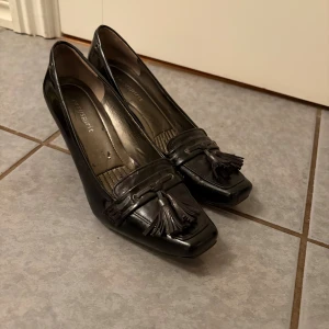 Svarta pumps med tofsar från Vagabond - Säljer ett par svarta klackskor från Easyspirit med dekorativa tofsar framtill och fyrkantig tå. Skorna har en låg klack och är tillverkade i blankt skinn. Perfekta för dig som vill ha en klassisk men ändå unik look.