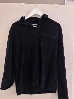 Svart manchestertröja från East West - Mjuk svart tröja från East West i manchester med relaxed fit och en stor bröstficka. Tröjan har krage, v-ringad öppning och långa ärmar. Perfekt för dig som gillar en chill och stilren look.