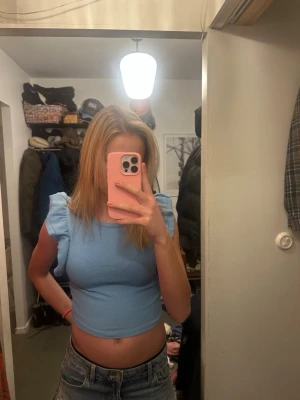 Ljusblå croppad topp med volangärmar - Säljer en ljusblå croppad topp med volangdetaljer på ärmarna. Toppen är ärmlös och har en rund halsringning. Materialet är ribbat och stretchigt, vilket gör den bekväm och snygg till jeans eller kjol. Säljer pga att det är för litet