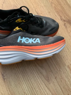 Svarta Hoka sneakers med orange sula - Säljer ett par snygga Hoka sneakers i svart med ljusblå logga och en riktigt chunky sula i vitt och orange. Skorna har snörning och ventilerande meshmaterial, perfekta för dig som gillar att sticka ut på löprundan.