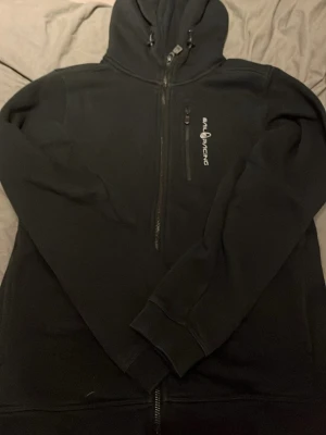 Svart hoodie från Sail Racing - Svart hoodie med dragkedja från Sail Racing. Tröjan har huva med dragsko, bröstficka med dragkedja och tryckt logga på vänster sida. I storlek 170. Den är använd ungefär fyra gånger för att den blev för liten.