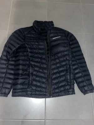 Svart dunjacka från Peak Performance - Snygg svart dunjacka från Peak Performance med quiltad design och dragkedja framtill. Jackan har hög krage och är perfekt för kalla dagar. Klassisk och stilren modell som funkar till det mesta.