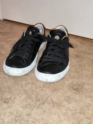 Svarta sneakers från Moncler - Svarta sneakers från Moncler med klassisk låg profil och rund tå. Skorna har svarta skosnören, vit sula och Moncler-logga på plösen. Tillverkade i skinn för en stilren look. Priset är ej hugget i sten!
