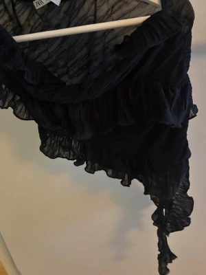 Svart volang topp från Zara - Säljer en svart, transparent topp från Zara med volangdetaljer och omlott-design. Toppen har ett axelband och är oneshoulder, vilket ger en unik och trendig look. Perfekt för dig som vill sticka ut med en cool och elegant stil.