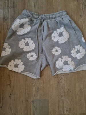 Grå shorts med moln från Denim Tears - Säljer ett par grå shorts från Denim Tears med vita molnliknande tryck över hela plagget. Shortsen har resårmidja med snörning och är tillverkade i mjuk bomull, perfekta för chill dagar. Snygg och unik design som sticker ut.