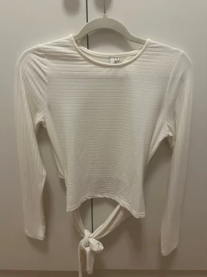 Vit ribbad långärmad topp med knyt i ryggen - Säljer en vit ribbad långärmad topp i mjukt stretchigt material. Toppen har rund halsringning och cool knytning baktill som ger en croppad look. Perfekt till jeans eller kjol för en trendig och enkel stil