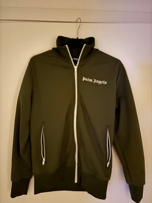 Palm Angels grön zip hoodie - Säljer en grön zip hoodie från Palm Angels med vit dragkedja och vita detaljer på ärmarna. Tröjan har hög krage, två fickor med dragkedja och logga på bröstet. Perfekt för dig som gillar streetwear och vill ha en clean look.