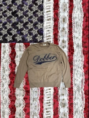 Tvärfet stickad Dobbersweater🌊 - Beige långärmad tröja från Dobber med rund halsringning och stort blått broderat tryck på bröstet. Tröjan har ribbade muddar vid ärmslut och nederkant. Perfekt för en avslappnad och stilren look.