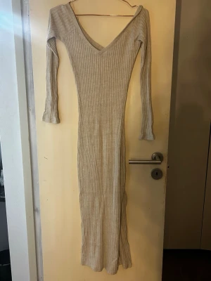 Beige ribbad långklänning - Långklänning i beige ribbat material med v-ringning och lå sopassform. Klänningen har långa ärmar och är figurnära, perfekt för en stilren och enkel look. Passar till många olika tillfällen och är lätt att matcha med accessoarer.
