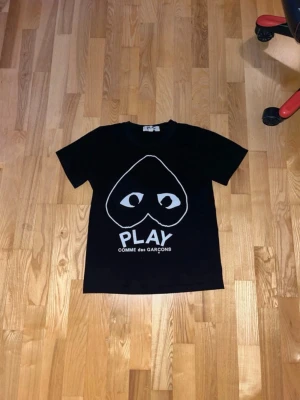 Svart Comme des Garçons Play t-shirt - Svart t-shirt från Comme des Garçons Play med det ikoniska hjärtat med ögon tryckt framtill och texten 'PLAY'. Klassisk passform och korta ärmar, perfekt för en stilren och trendig look.