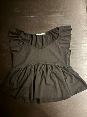 Svart volangtopp från 157 - Säljer en svart topp från 157 med volangdetaljer och korta ärmar. Toppen har en lätt ribbad struktur och en lös passform nedtill. Perfekt att styla med jeans eller kjol för en trendig look.