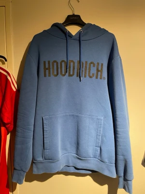 Blå hoodie från Hoodrich - Snygg blå hoodie från Hoodrich med stor svart logga på bröstet. Hoodien har huva med dragsko och en stor magficka framtill. Perfekt för en avslappnad och trendig stil. Tillverkad i mjukt material som känns skönt mot huden.