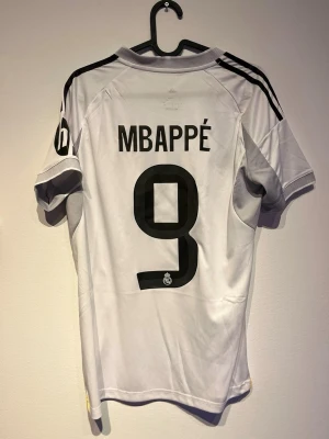 Real Madrid Mbappé hemmatröja - Säljer en vit Real Madrid matchtröja från Adidas med Mbappé och nummer 9 på ryggen. Tröjan har svarta detaljer, gula inslag och klubbmärke på bröstet. Materialet är lätt och andas, perfekt för fotbollsträning eller match.