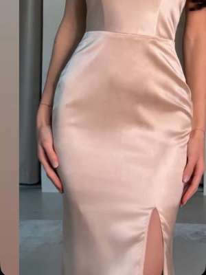 Champagnefärgad långklänning i satin - Elegant långklänning i satin med figurnära passform och slits framtill. Klänningen har en mjuk glansig yta och är ärmlös, perfekt för speciella tillfällen. Inköpt på Dressrom för 2500kr