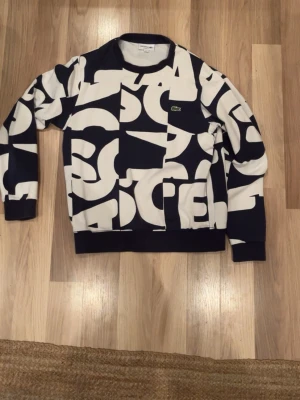 Lacoste tröja  - Säljer en riktigt snygg  sweatshirt från Lacoste i storlek M. Tröjan har ett stort grafiskt mönster och den klassiska gröna krokodilloggan på bröstet. Skön passform med rund hals och långa ärmar. Perfekt för dig som gillar statement-plagg!