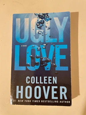Ugly Love av Colleen Hoover - En engelskspråkig pocketbok med titeln 'Ugly Love' av Colleen Hoover. Omslaget är blått med stora bokstäver och bubblor som ger en vatteneffekt. Perfekt för dig som gillar romantiska och känslosamma berättelser.