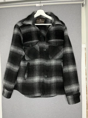 Svartgrå rutig jacka Oscar Jacobson - Snygg svart och grå rutig jacka från Oscar Jacobson. Jackan har två bröstfickor med lock och knappar, samt två sidofickor. Klassisk skjortjackemodell med krage och knäppning framtill. Tillverkad i ett mjukt, ull-liknande material.
